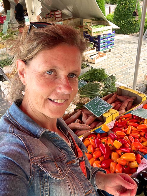 Béatrice, la fondatrice de Femmes en Harmonie, sur un marché local devant l'état d'un producteur de fruits et légumes du mois de juillet.