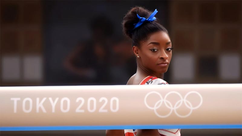 Simone se tient derrière la poutre marquée 'Tokyo 2020' avec les cinq anneaux olympiques, observant cette poutre avec une concentration intense.
