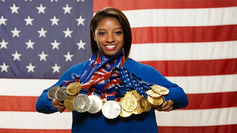 Simone Biles, souriante et triomphante, montre fièrement ses nombreuses médailles autour de son cou, avec un immense drapeau américain en arrière-plan. Cette image capture le summum de sa carrière exceptionnelle, incluant 25 médailles mondiales, dont 19 en or.