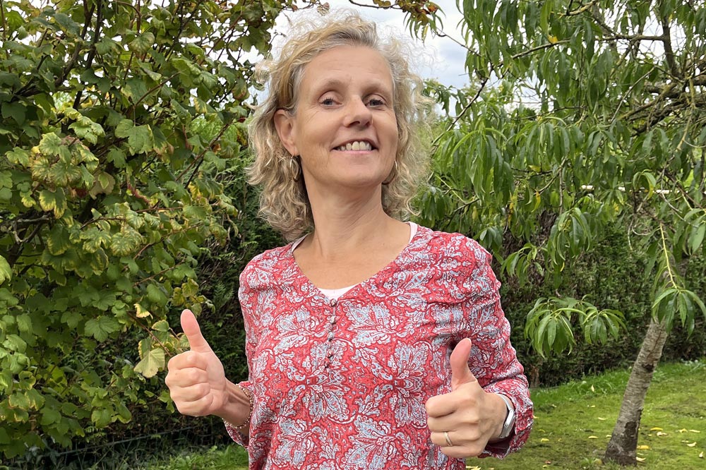 Béa, fondatrice de Femmes en Harmonie, dans son jardin avec un grand sourire et les pouces levés, portant un chemisier coloré à fleurs, encourageant les femmes à retrouver la motivation et à cultiver un bien-être durable.