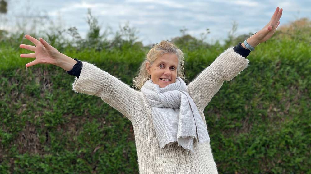 Béa, fondatrice de Femmes en Harmonie, souriante et bras ouverts dans son jardin, pour conclure avec motivation et inspiration cet article sur la peur de vieillir. Béa, à 55 ans, symbolise la joie, la confiance et la sérénité face au temps qui passe.