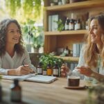 Femme d’environ 45 ou 50 ans en consultation dans un cabinet de naturopathie, face à une naturopathe souriante. Sur le bureau en bois clair, des plantes, des compléments alimentaires, des huiles essentielles et une tisane évoquent les méthodes naturelles. L’ambiance zen et chaleureuse reflète un accompagnement bienveillant pour mieux vivre la ménopause et retrouver l’équilibre.