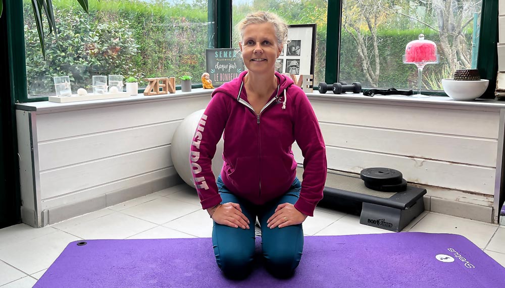 Béa assise sur un tapis de yoga violet, souriante après une séance d’exercices. La pratique régulière d’exercices ciblés aide à obtenir un ventre plat, améliorer la posture et booster la confiance en soi, tout en réduisant le stress grâce à la libération d’endorphines.