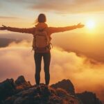 Femme confiante d'environ 50 ans, debout au sommet d’une montagne au lever du soleil, les bras ouverts vers l’avenir. Son expression sereine illustre l’impact du coaching mental pour se libérer du stress, renforcer son mindset positif et retrouver confiance en soi.