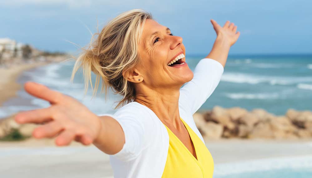 Femme épanouie souriante au bord de la mer, symbole de renaissance et de mieux-être après avoir traversé les symptômes de la ménopause : anxiété, fatigue, bouffées de chaleur, troubles du sommeil.